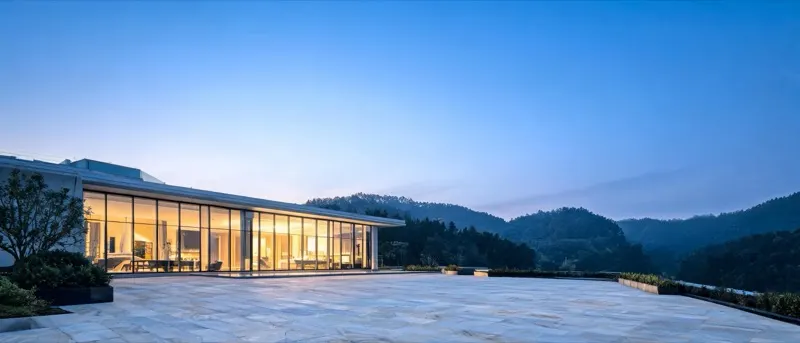 Moderne Villa mit beleuchteter Glasfassade zur blauen Stunde, dahinter Hügel (KI-generiertes Bild)